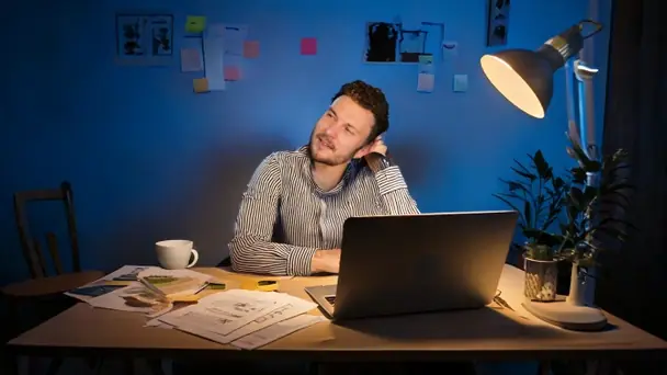A man at a home desk at night, laptop open and notes on the wall, leaning back with a hand behind his neck