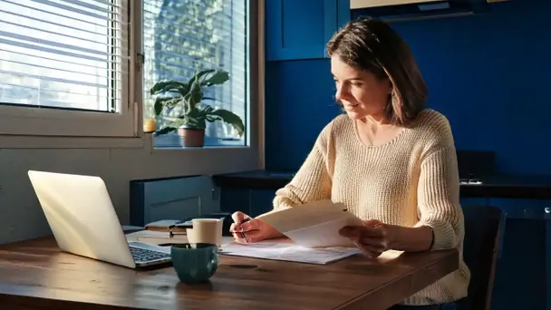 A woman at a kitchen table with a laptop and a folder of paperwork, reading in daylight