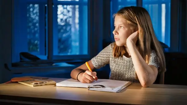 A girl at a table in the evening with an open notebook and a pencil, looking out of the window