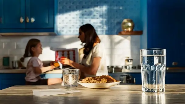 A kitchen counter early in the morning with a glass of water and a wall calendar, a parent and a school-age child eating breakfast softly out of focus behind
