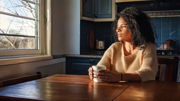 A woman sitting at a kitchen table with a mug, looking out of the window in morning light