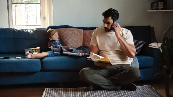 A father on the phone sitting beside a sofa while a young child looks at a book next to him