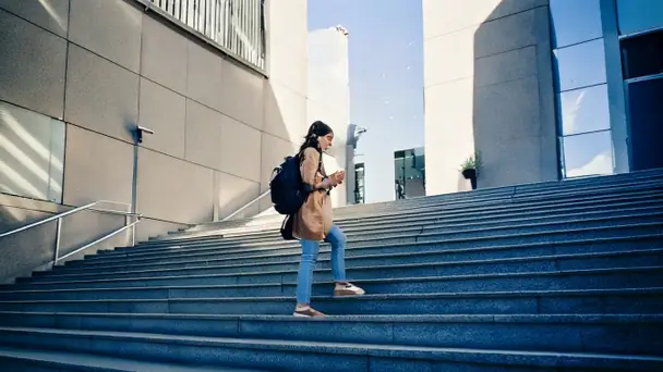 A student with a backpack walking up a flight of concrete steps outside a building, looking at a phone