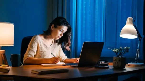 A woman at a small table in the evening with a laptop open, writing on a notepad by lamplight