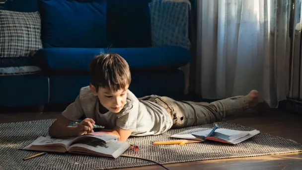 A boy lying on a rug reading a large open book, with a worksheet and pencil set aside nearby